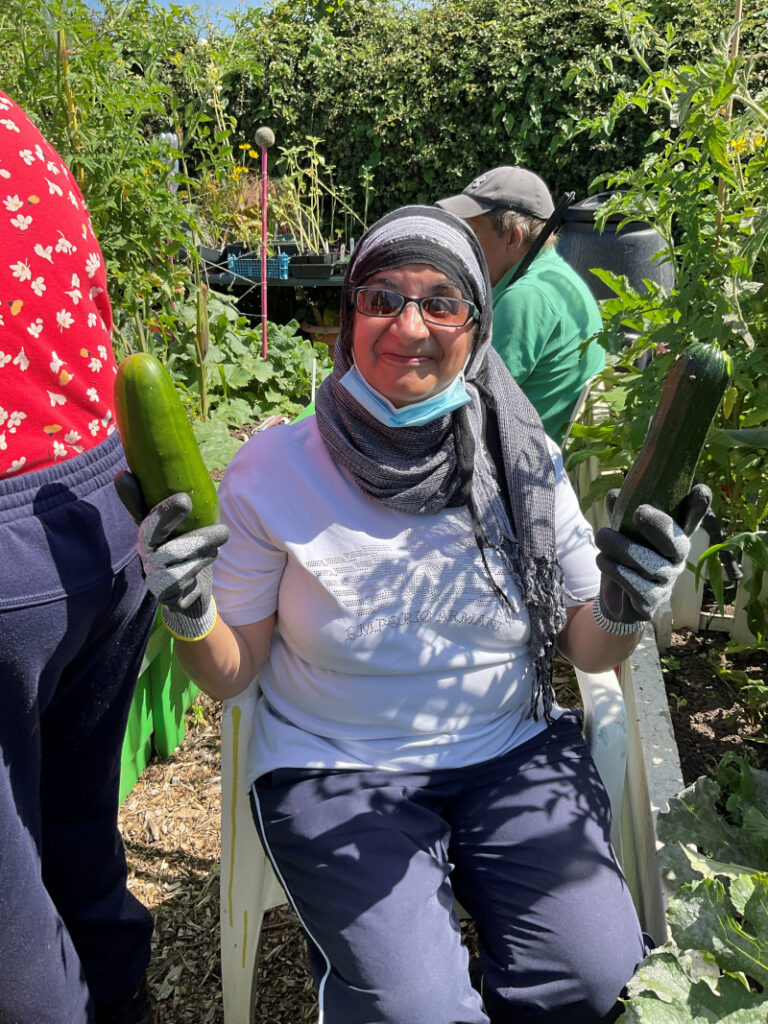 A seated woman in a hijab at the allotment holding up a cucumber and courgette