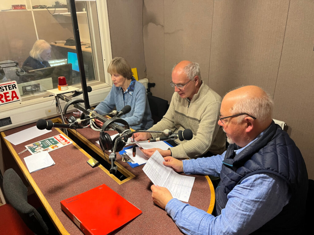 Three people seated around a table in a recording studio and a sound engineer through glass