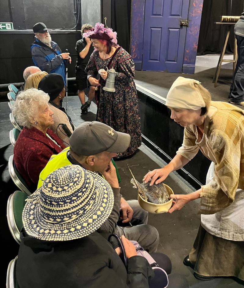 Visually impaired people seated in a theatre with actors demonstrating props and costumes