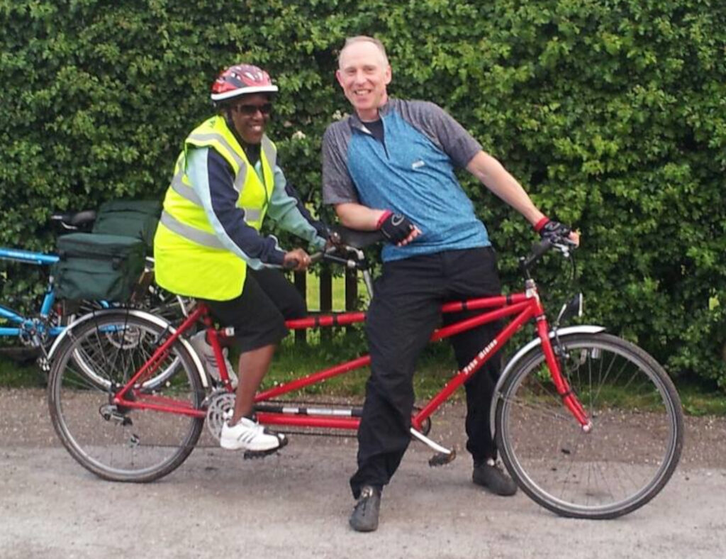 A visually impaired ‘stoker’ on the rear seat of a tandem bike, with a sighted ‘pilot’ leaning alongside